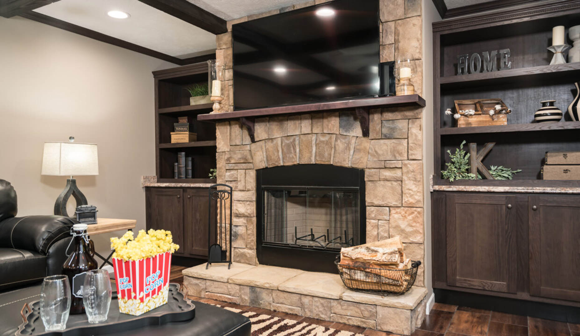 A cozy living room features a stone fireplace with a large TV above it, flanked by dark wood shelves. A leather sofa and a table with a popcorn bucket create a warm, inviting atmosphere.