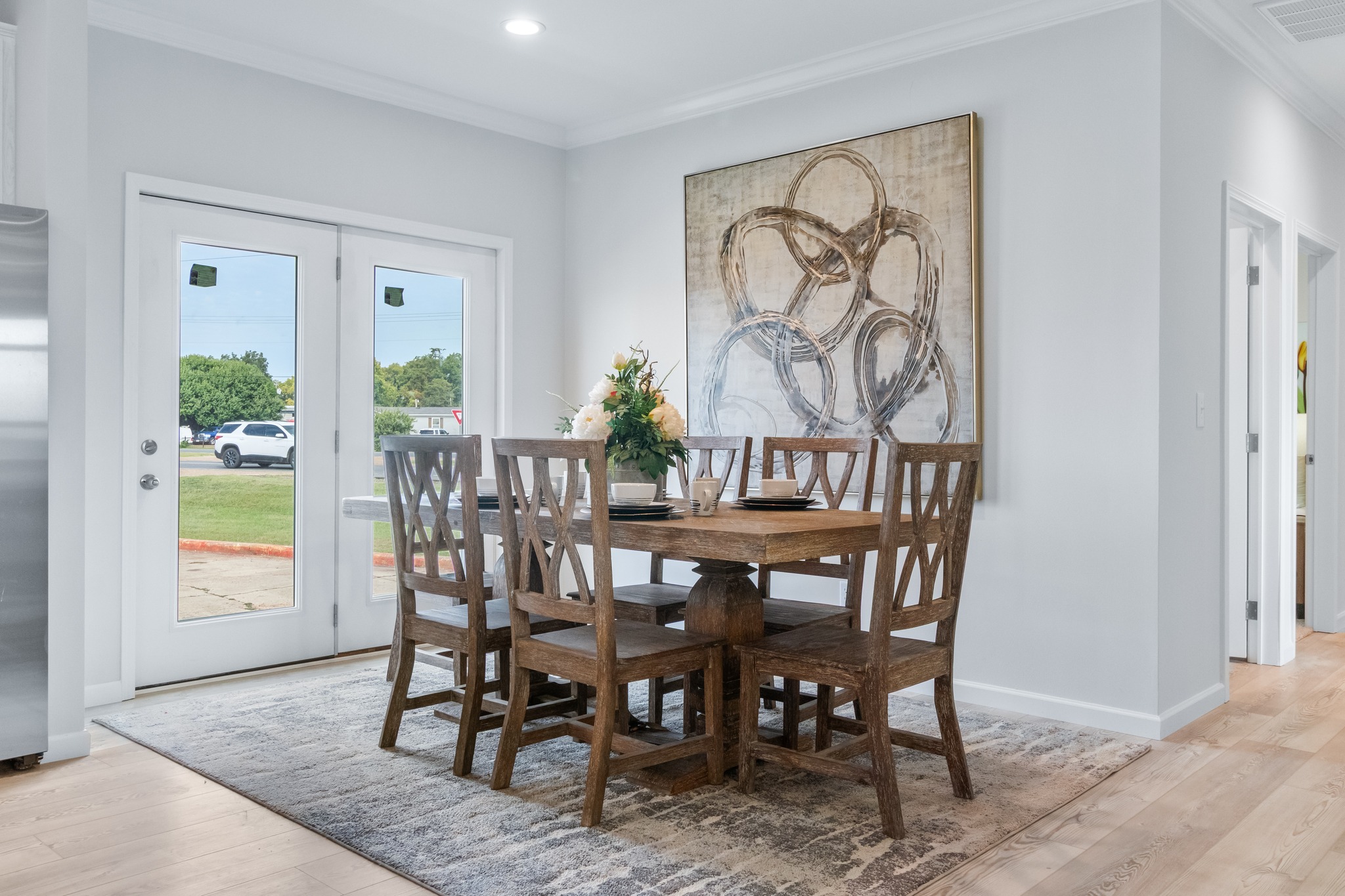 Dining room with a wooden table and six chairs on a patterned rug, large abstract art on the wall, and glass doors leading outside. Bright and inviting atmosphere.