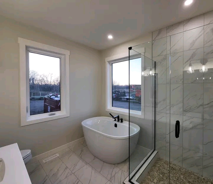Modern bathroom with a freestanding white bathtub, marble-tiled shower, and two large windows. Soft lighting creates a serene atmosphere.