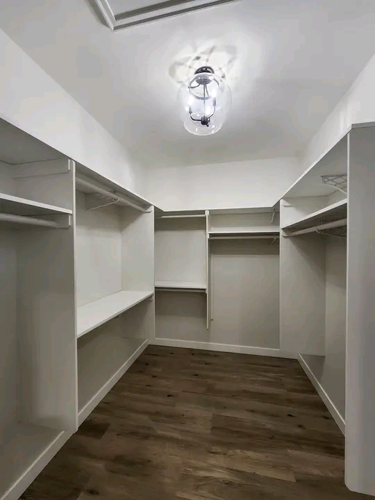 Spacious, empty walk-in closet with white shelves and hanging rods on both sides. Wooden floor and a ceiling light create a clean, minimalist feel.