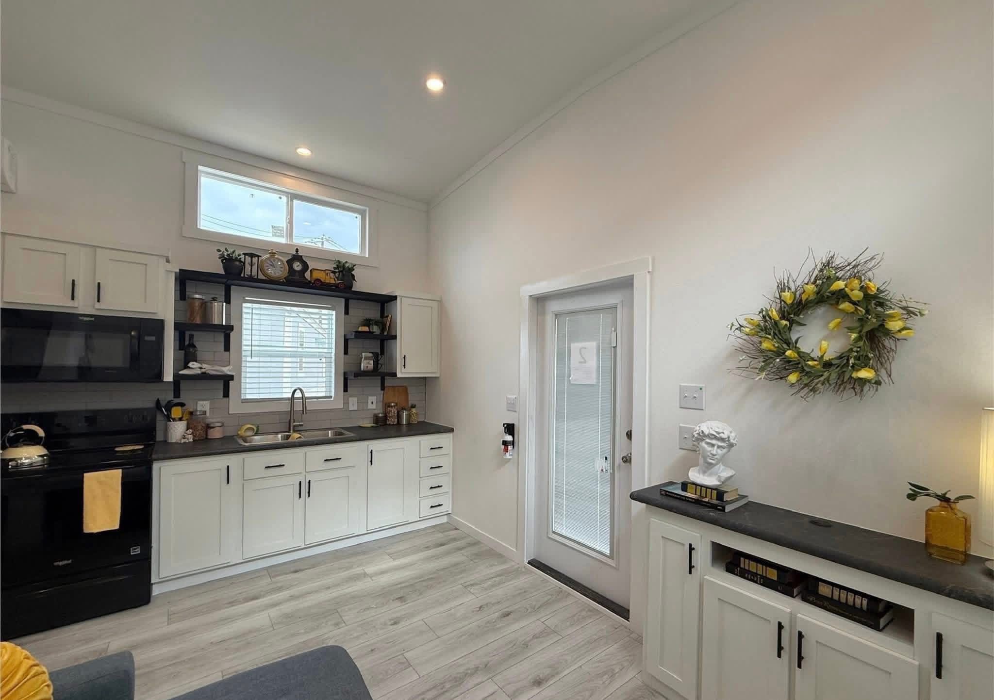 Modern kitchen with white cabinets, black countertop, and appliances. A window and door add natural light. A lemon wreath decorates a wall.