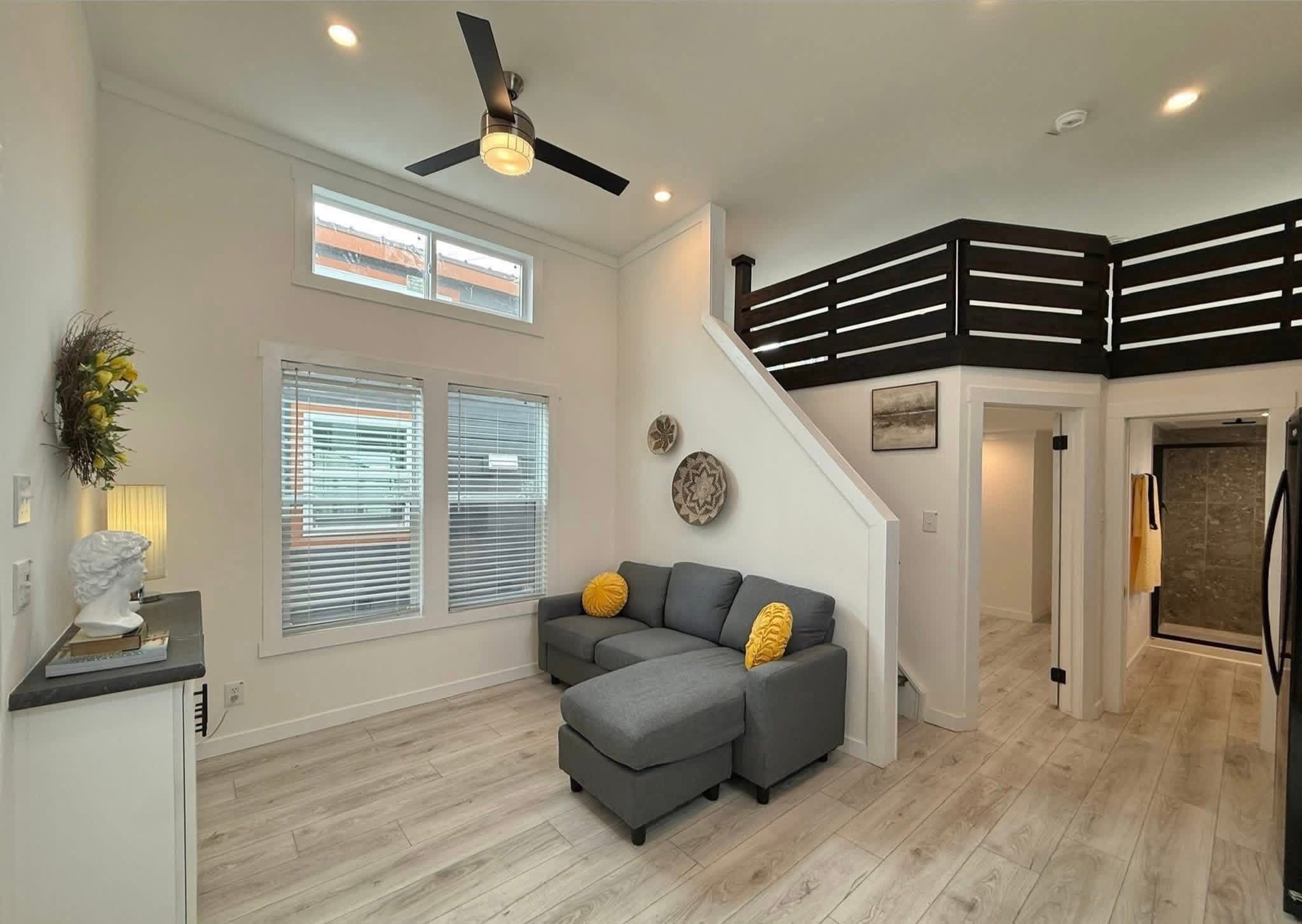 Modern living room with light wood flooring, gray sectional sofa, and yellow pillows. Black ceiling fan above, stairs lead to loft. Bright and cozy.