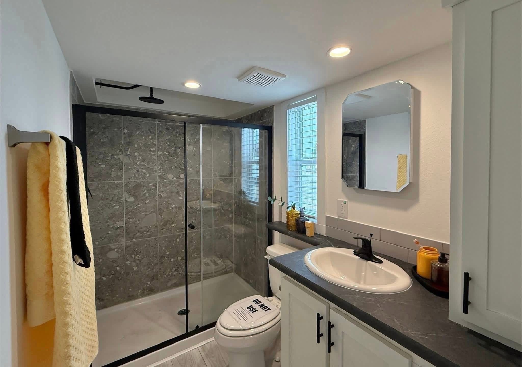 Modern bathroom with gray tile shower, glass door, and black fixtures. White cabinets, round mirror, and colorful towels create a sleek, cozy vibe.
