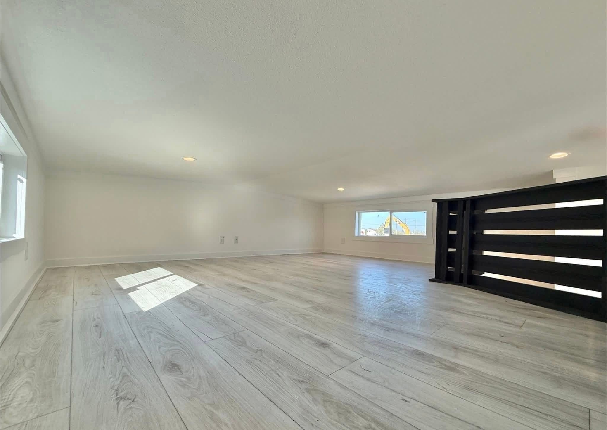Bright, minimalist attic space with light wood flooring and white walls. Two small windows let in natural light, creating a serene, airy atmosphere.