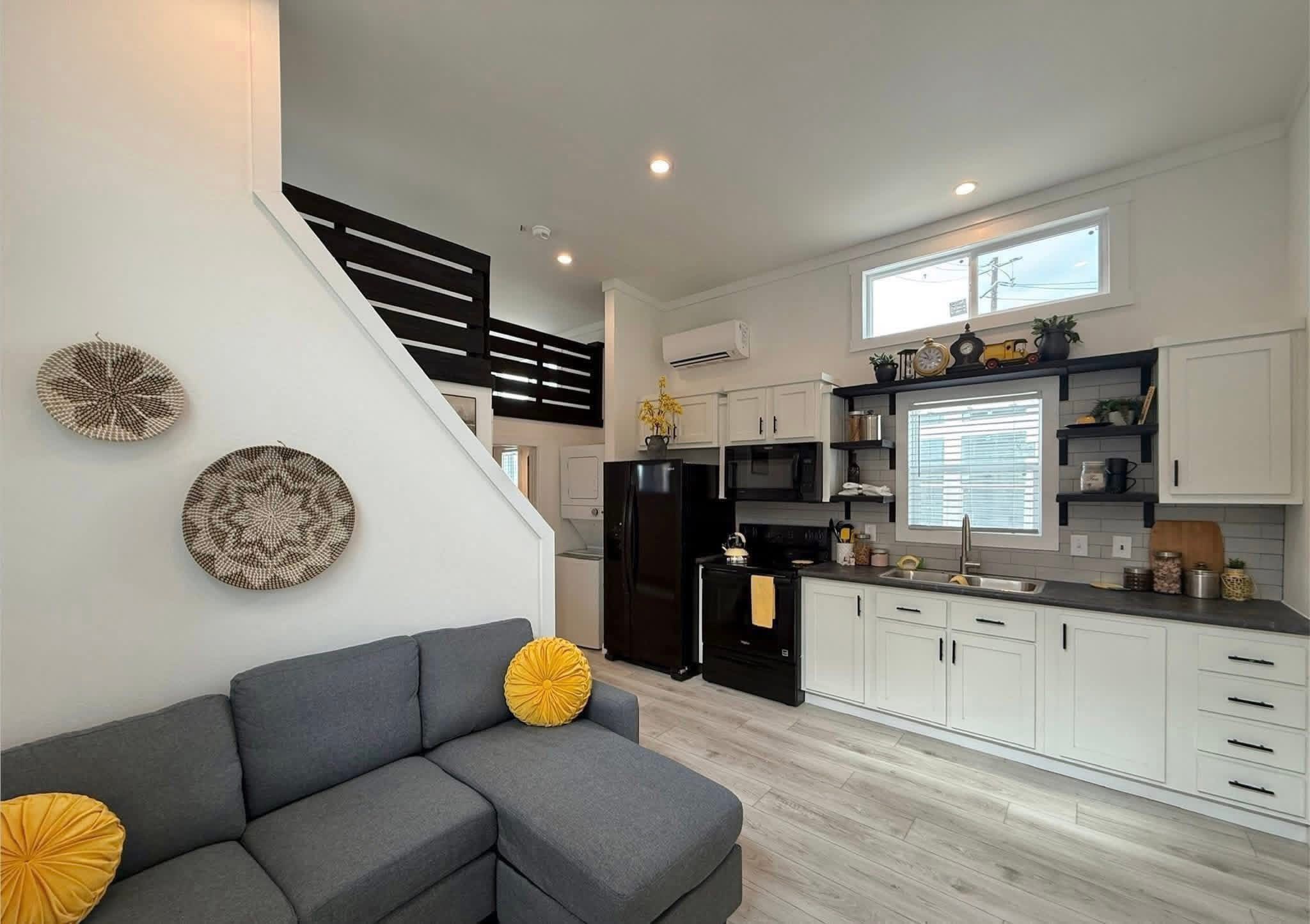 Modern tiny home interior with gray sectional sofa adorned by yellow pillows. White kitchen features black appliances and minimalist decor. Cozy, bright ambiance.