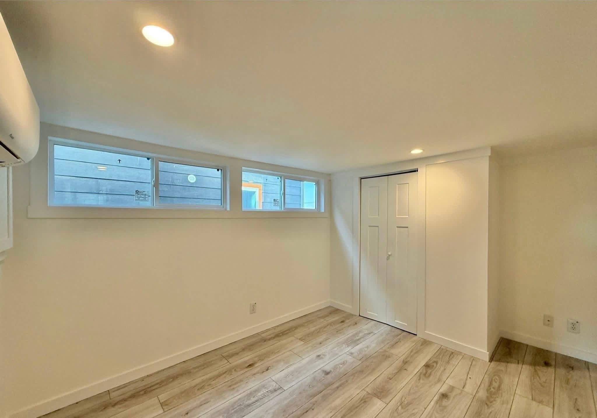 Minimalist room with light wood flooring, an air conditioning unit on the left wall, high horizontal windows, and a small white closet door.