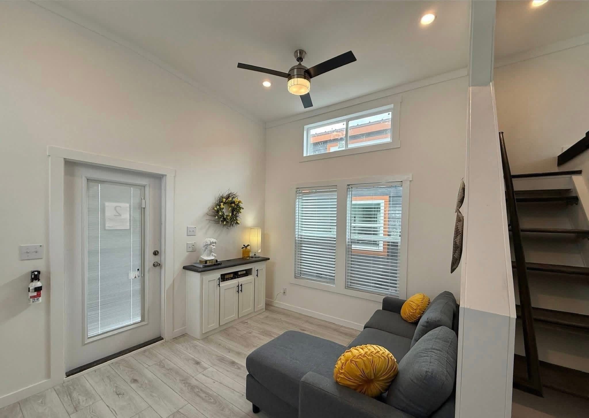 Bright living room with gray sofa and yellow cushions under tall windows. A modern ceiling fan hangs above, and a staircase leads to an upper level.