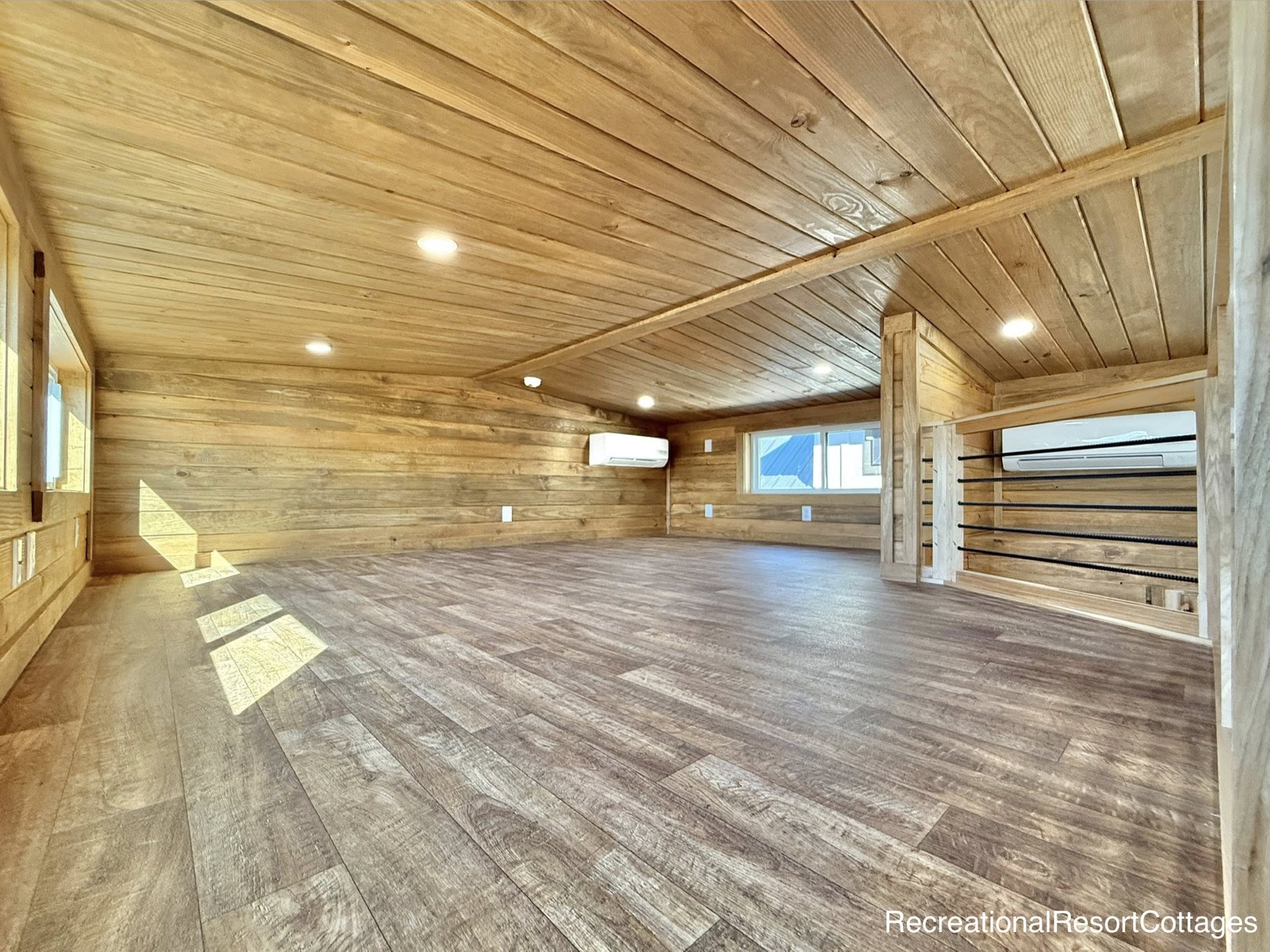 Spacious wooden loft with warm lighting, featuring wooden ceiling and floor, large windows on the left, and compact air conditioning unit on the wall.