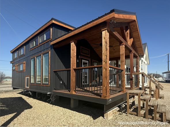 A sleek black tiny house with a gabled roof and wooden accents, featuring large windows and a cozy porch under a clear blue sky, conveying modern comfort.