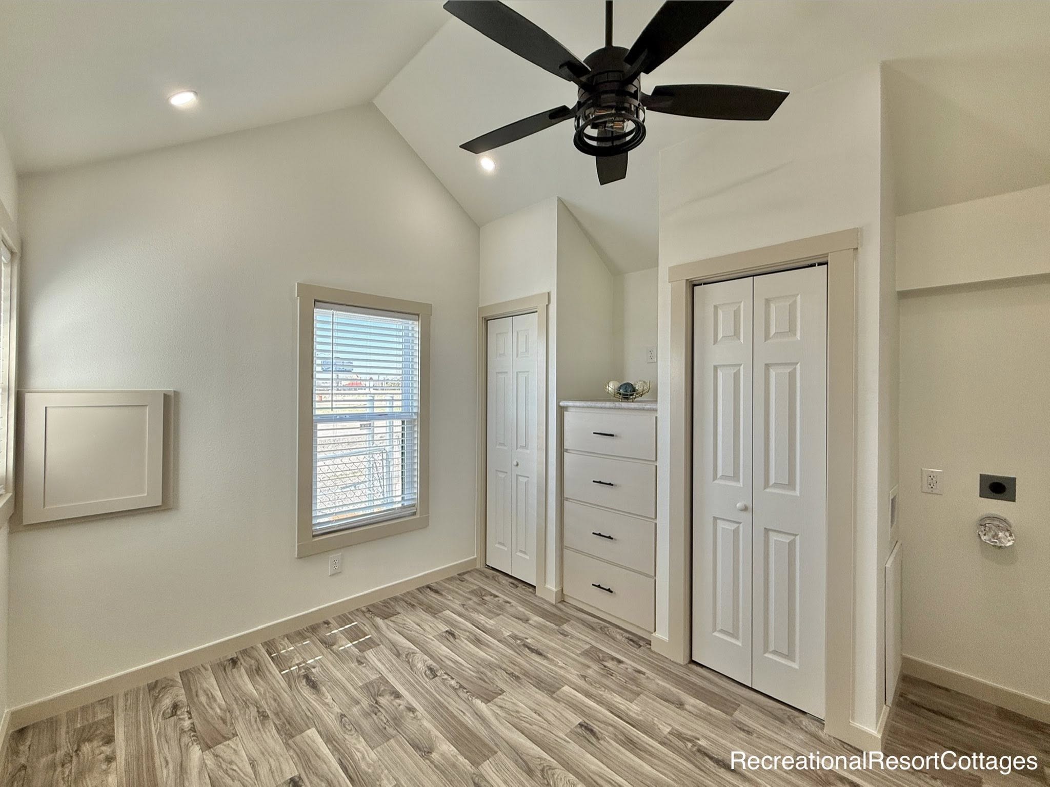 Bright room with vaulted ceiling, featuring wood-style flooring, a ceiling fan, a window with blinds, white closet doors, and built-in drawers.