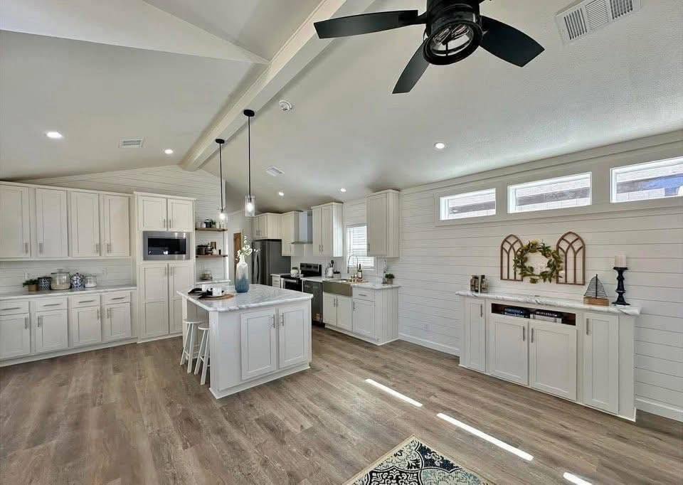 Spacious kitchen with white cabinetry, a central island, pendant lights, wood flooring, ceiling fan, and natural light from high windows, creating an airy feel.
