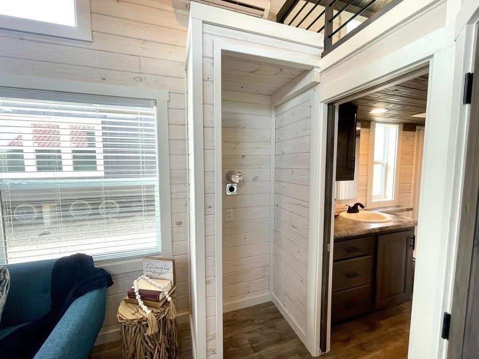 Cozy tiny house interior featuring light wood paneling, a small bathroom with a sink, and a living area with a blue chair, stack of books, and window.