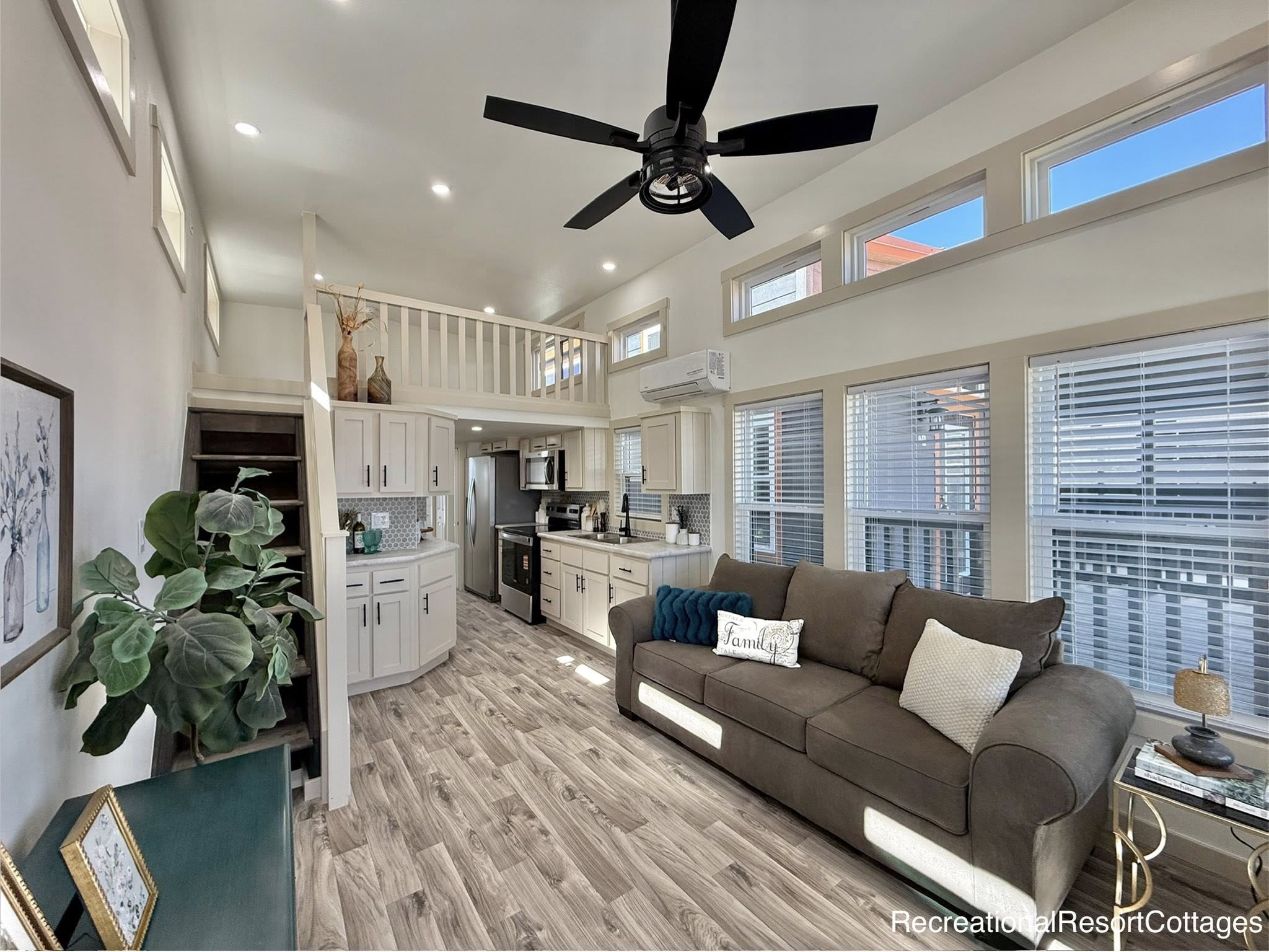 Bright tiny home interior with wood flooring, loft space, kitchen, and cozy sitting area. Large windows and ceiling fan enhance airy feel.