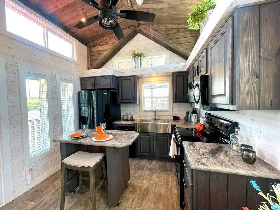 Cozy kitchen with dark wood cabinets, marble countertops, center island with stools, stainless steel appliances, and potted plants, under a vaulted ceiling.