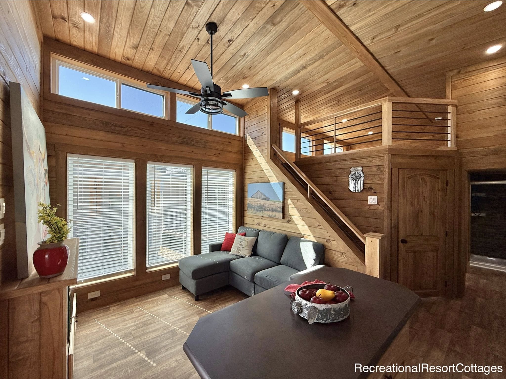 Cozy cabin interior with wood paneling, a gray L-shaped sofa, a loft, and large windows. A ceiling fan and natural light create warmth.