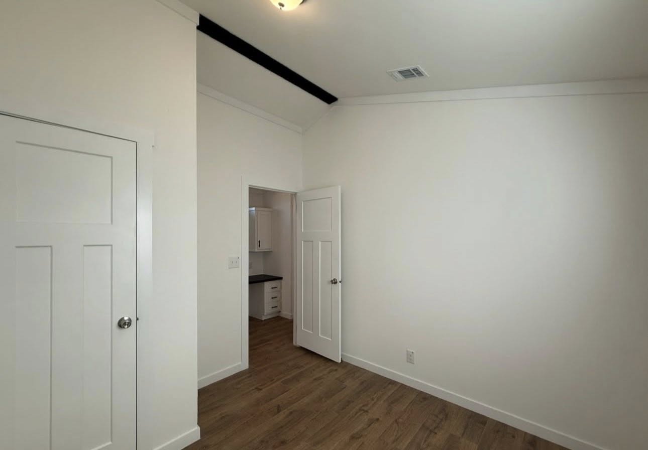 A clean, empty room with white walls and wood flooring. A door is slightly open, revealing a bright adjacent room and cabinets. The space feels modern and minimalistic.