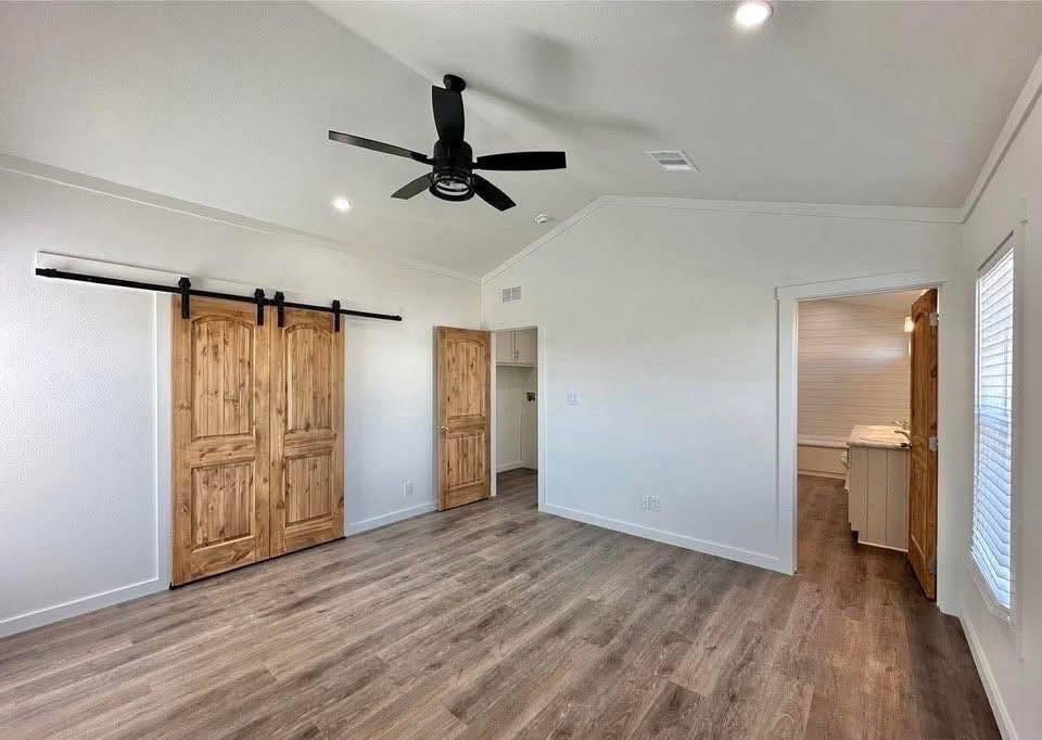 Spacious room with wooden flooring, sliding barn doors, and modern black ceiling fan. Adjacent room visible through an open door. Bright, minimalist feel.
