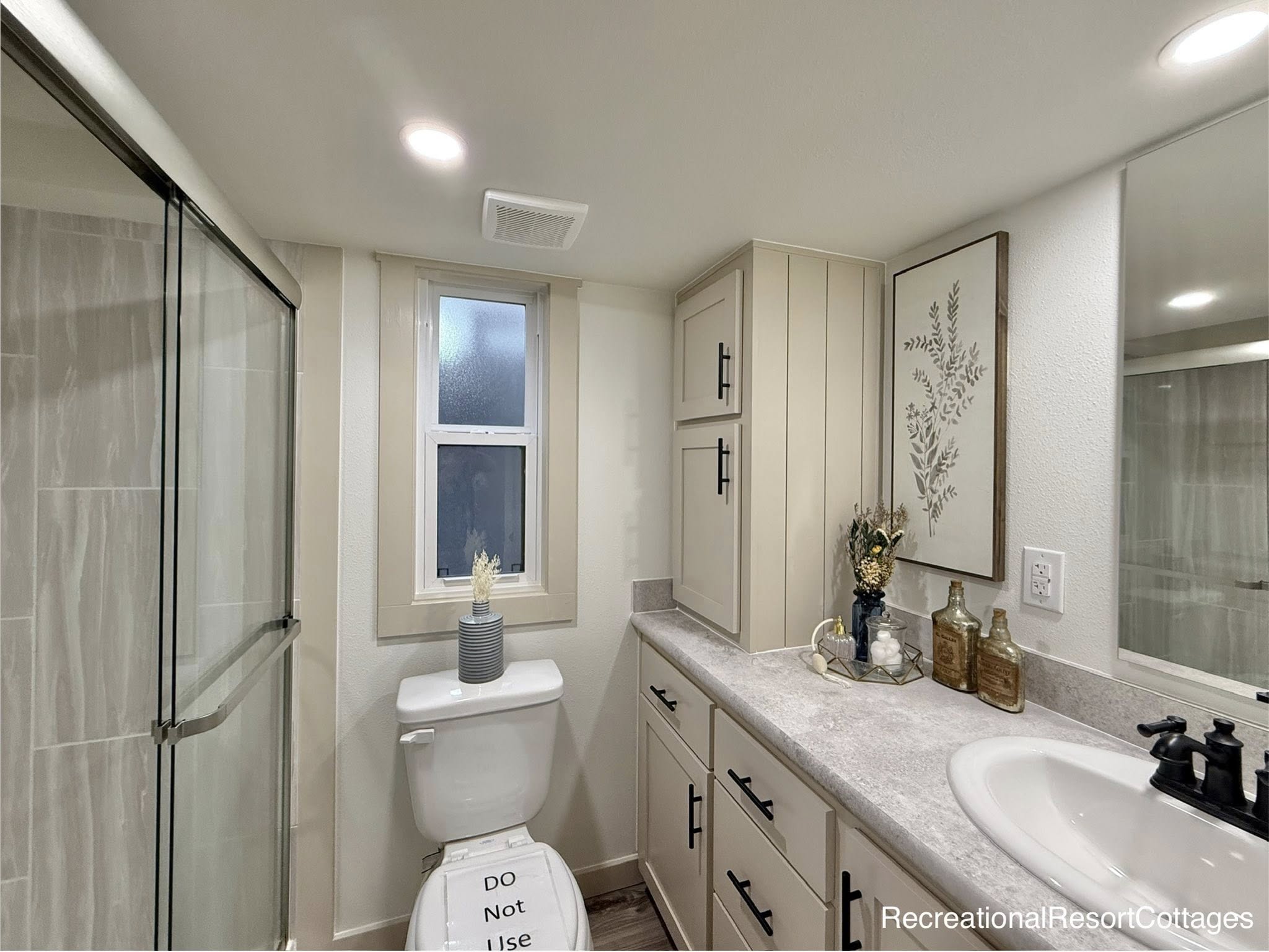 Modern bathroom with a glass shower, beige cabinets, and white countertop. Decorative items on the counter add elegance. Bright and clean atmosphere.