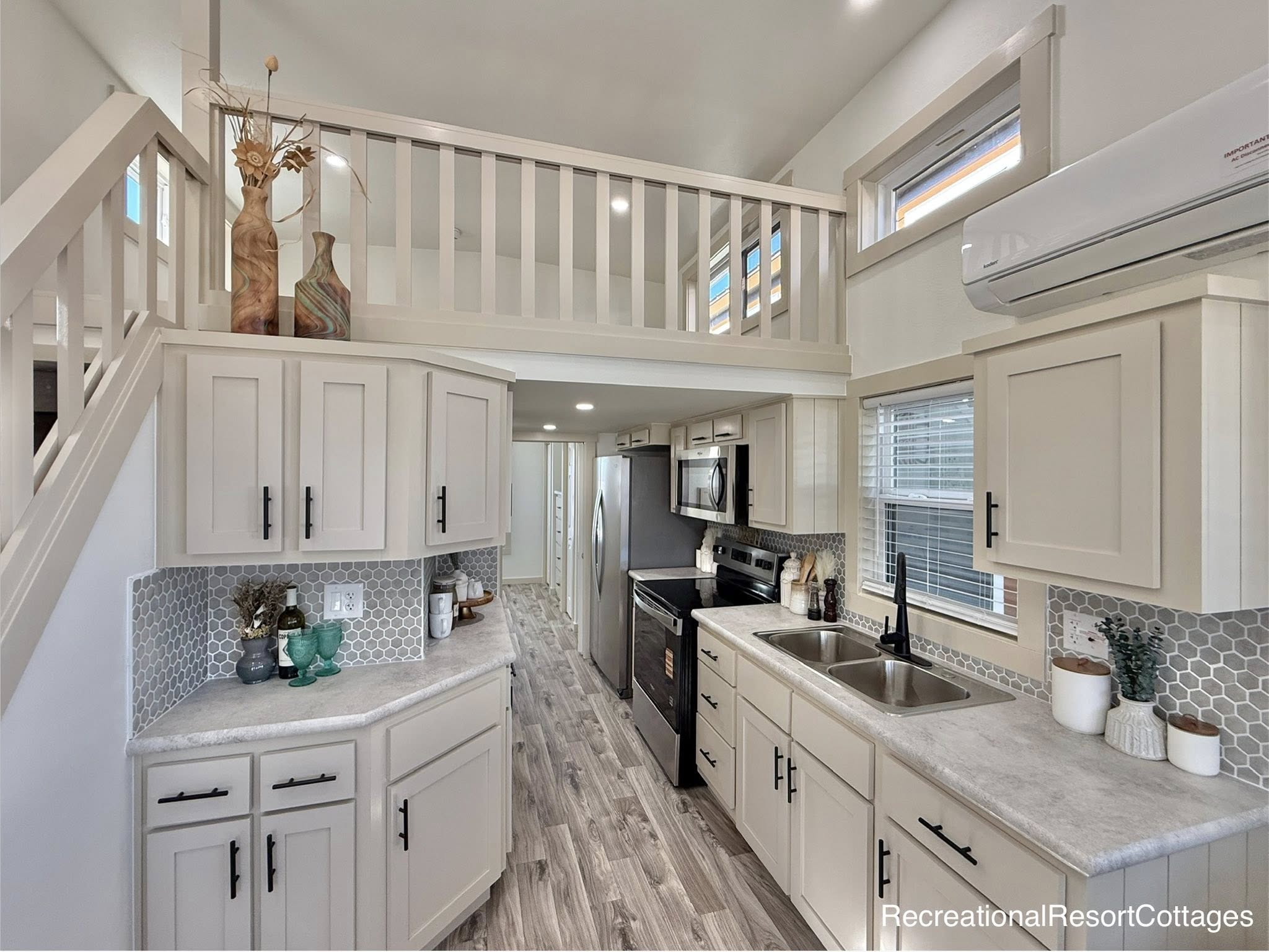 Compact modern kitchen with beige cabinets, stainless steel appliances, and light wooden flooring. An open loft and large windows provide airy brightness.