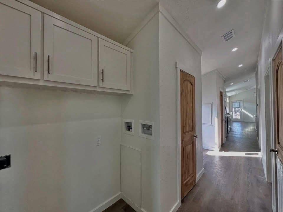 Hallway with wooden flooring, sunlight streaming in. White cabinets on the left, wooden doors, and visible kitchen area in the distance create a warm, inviting ambiance.
