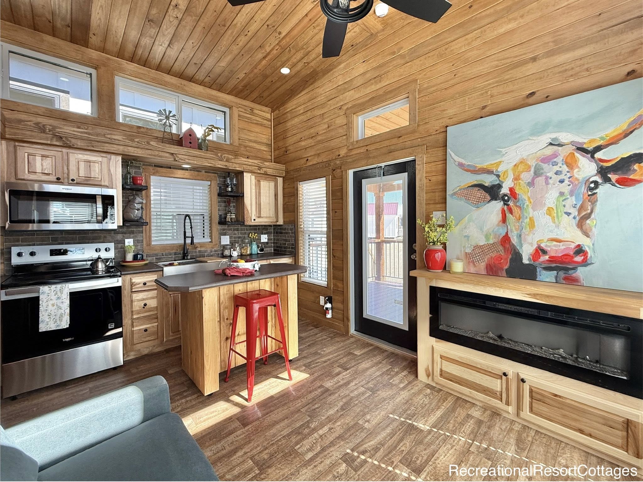 Cozy cabin kitchen with warm wood paneling, a colorful cow painting, modern appliances, red bar stool, and a bright, inviting atmosphere.