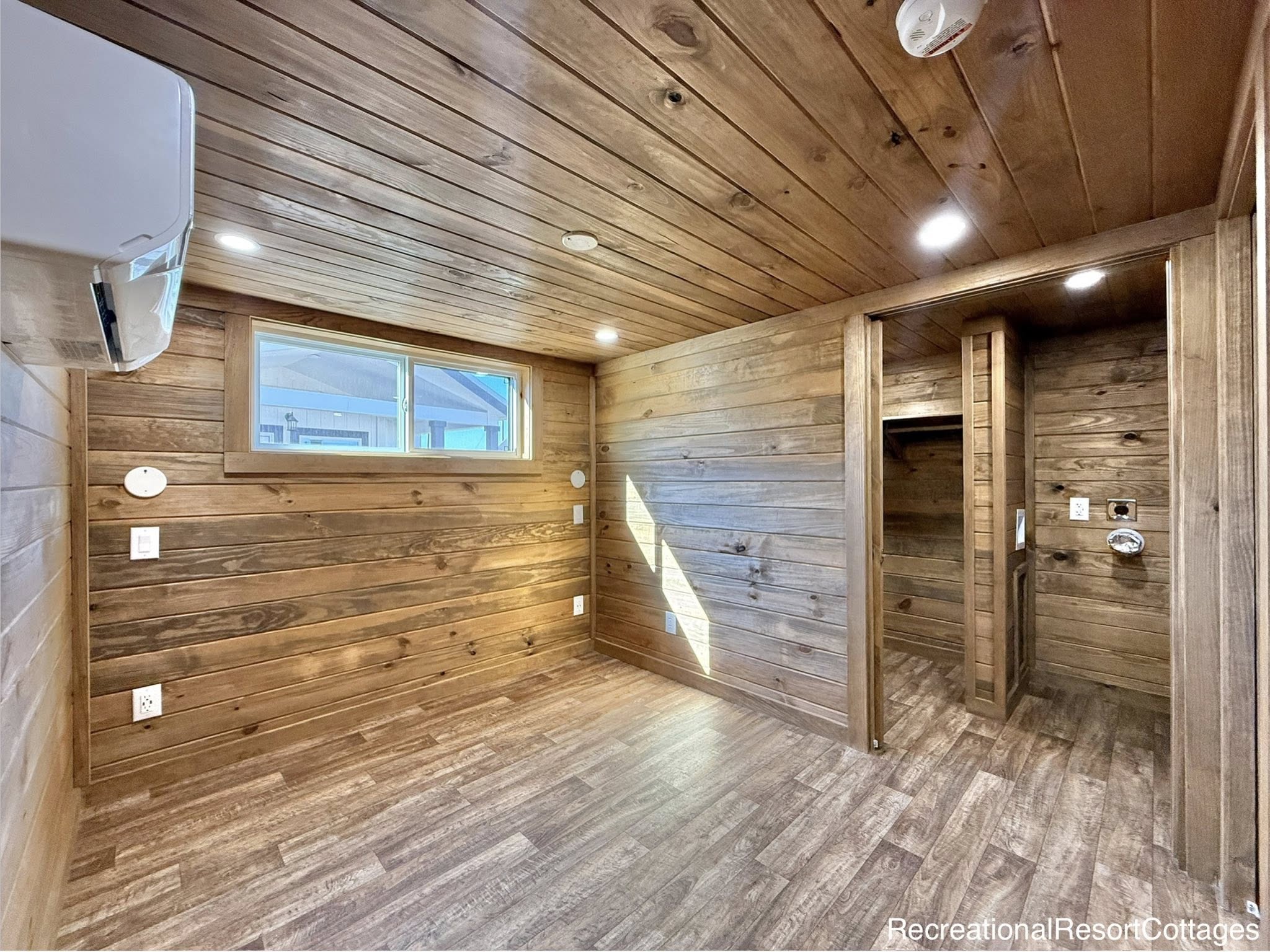 Compact wooden room with natural light from a horizontal window, wood-paneled walls and ceiling, recessed lighting, vinyl flooring, and an open closet space. Cozy and rustic ambiance.