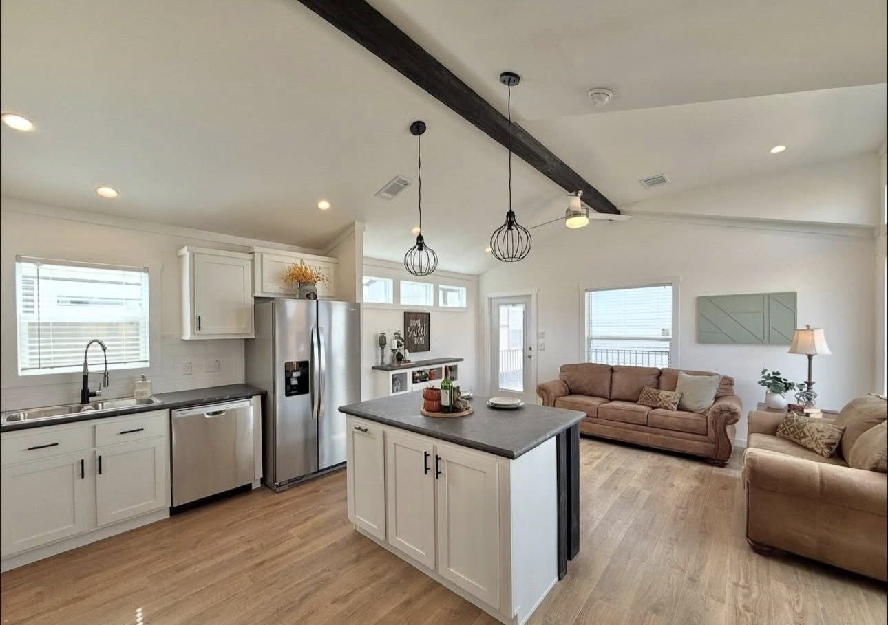 A modern, open-concept kitchen and living room with hardwood floors. The kitchen features white cabinets, stainless steel appliances, and a central island. Two beige sofas and a small table lamp create a cozy living space. Black pendant lights hang above, adding a contemporary touch.