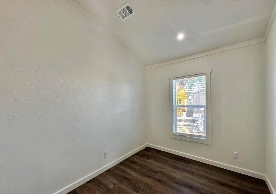 Small empty room with white walls and dark wood flooring. A single window shows a view of houses outside. The space feels bright and minimalistic.
