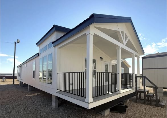 A modern white mobile home with large windows and a small covered porch, featuring black railings. It's set on gravel under a clear blue sky.