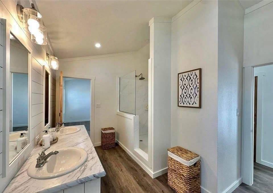 Bright modern bathroom with dual sinks on a marble countertop, elegant wall lights, walk-in shower, wicker baskets, hardwood floor, and framed art.
