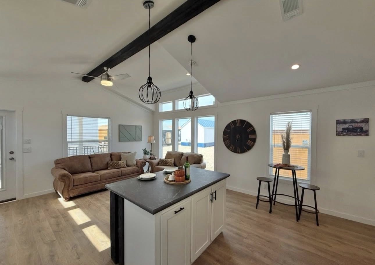 Spacious living room with vaulted ceiling, modern pendant lights, brown sofas, and a central island. Large windows illuminate the cozy decor.