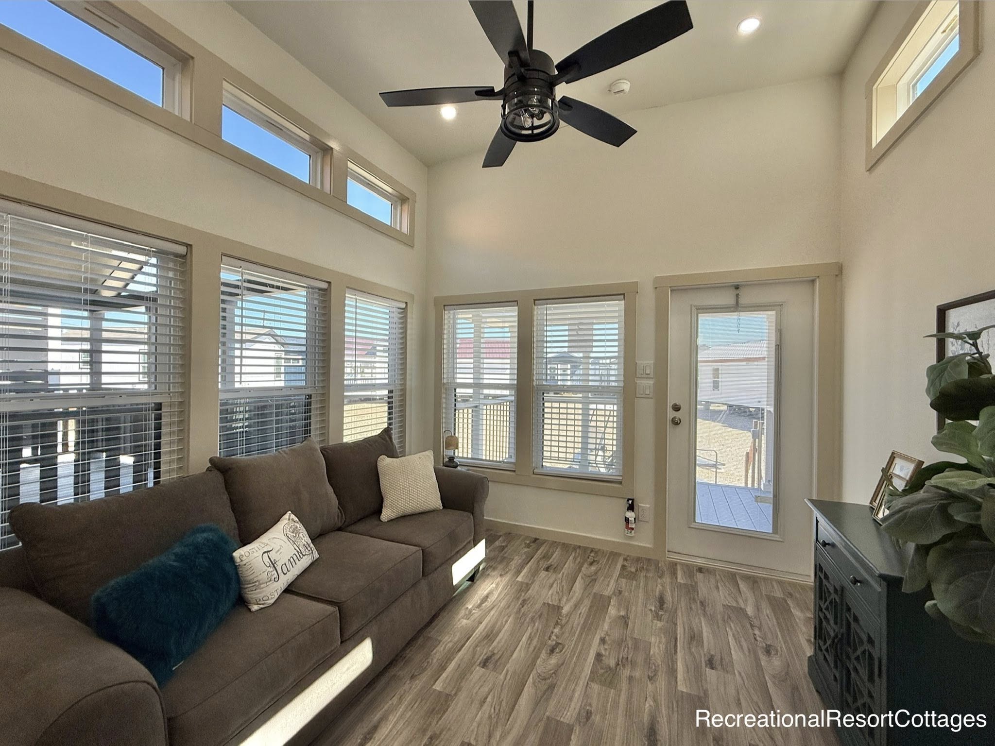Modern living room with high ceiling and large windows, featuring a brown sofa with decorative pillows, a ceiling fan, and wood-style flooring. Bright and airy.