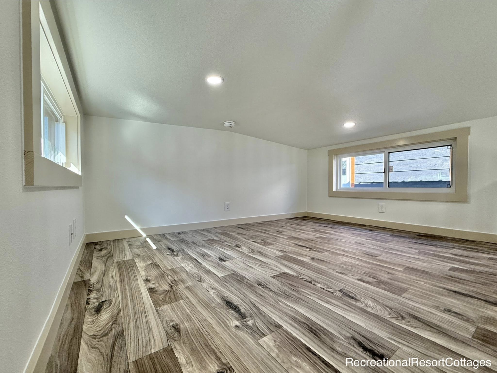 Spacious room with a sloped ceiling and light wood flooring. Two large windows let in natural light. The atmosphere is bright and airy.