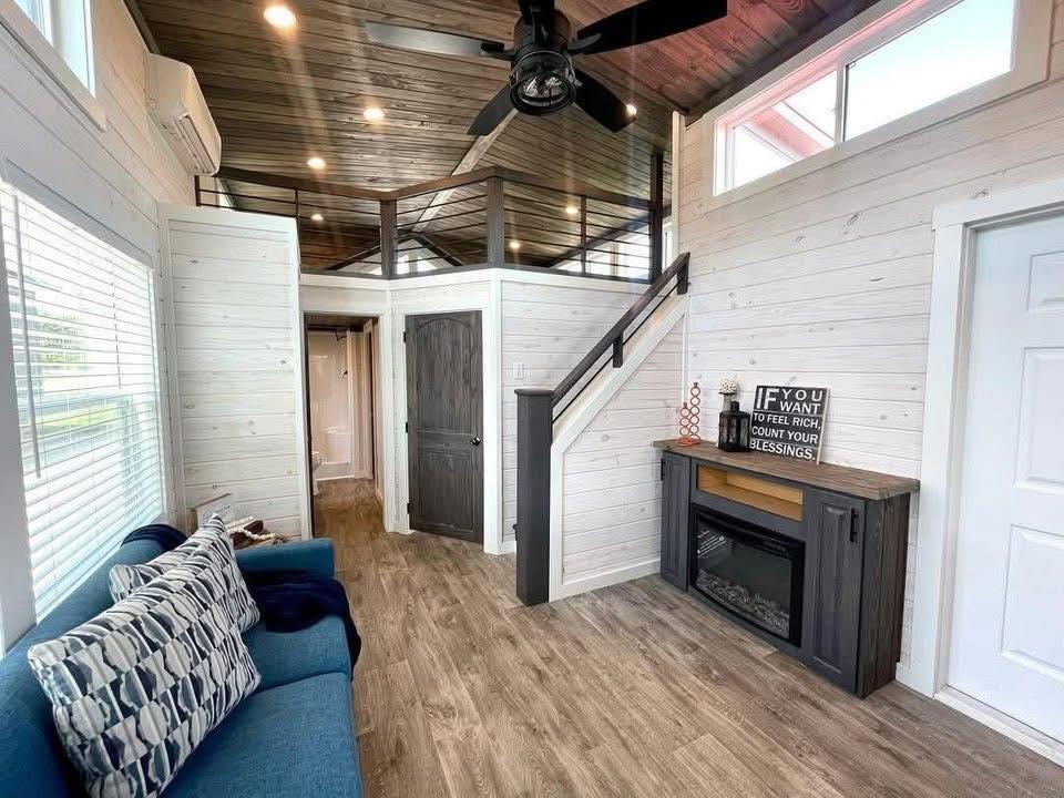 Cozy tiny house living room with white shiplap walls, dark wood ceiling, and light wood floors. Features a blue couch with patterned pillows, an electric fireplace with a motivational sign, and a loft accessed by a wooden staircase.