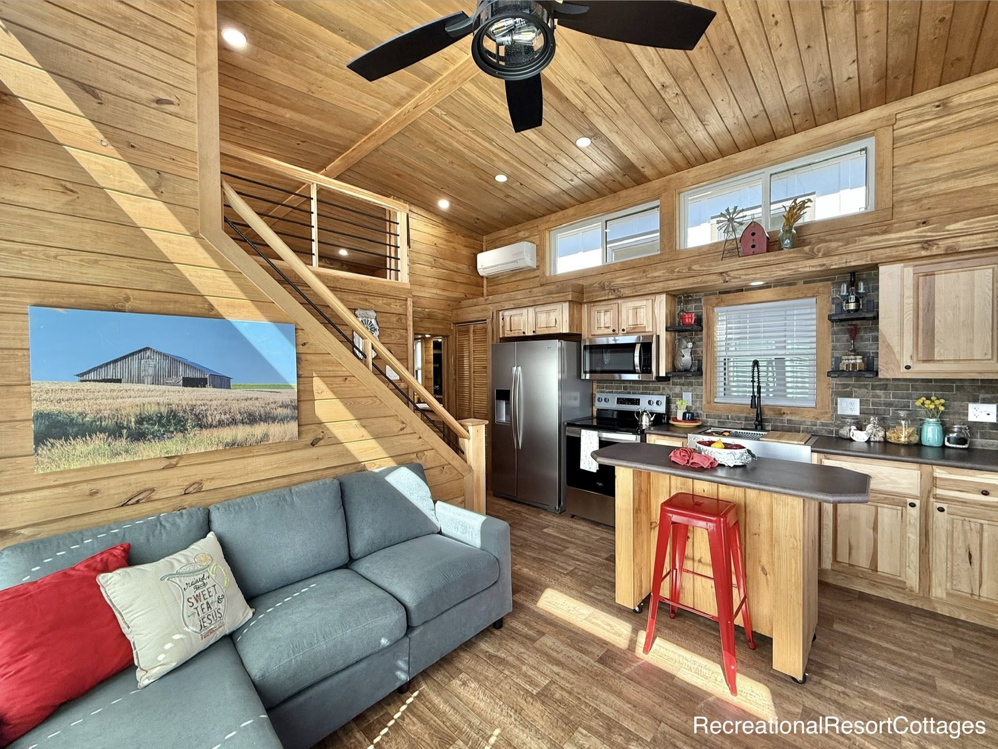Cozy wooden cabin interior with natural light. Features a gray couch, red accents, kitchen with stainless appliances, loft, and rustic decor.