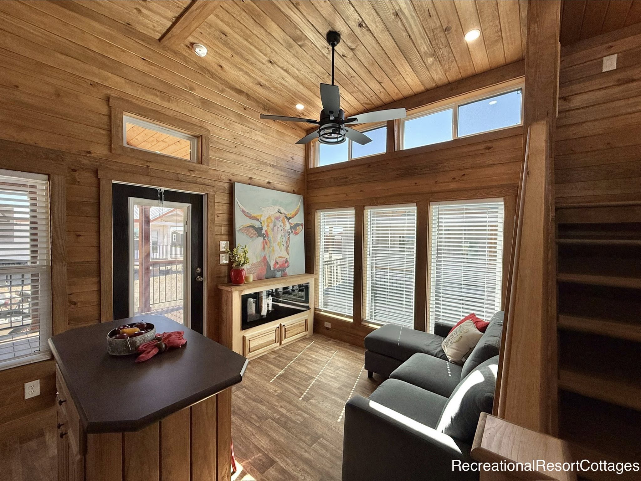 Cozy cabin interior with wooden walls and ceiling. Sunlit room features a gray sectional, cow artwork, kitchen island, and ceiling fan, exuding warmth.