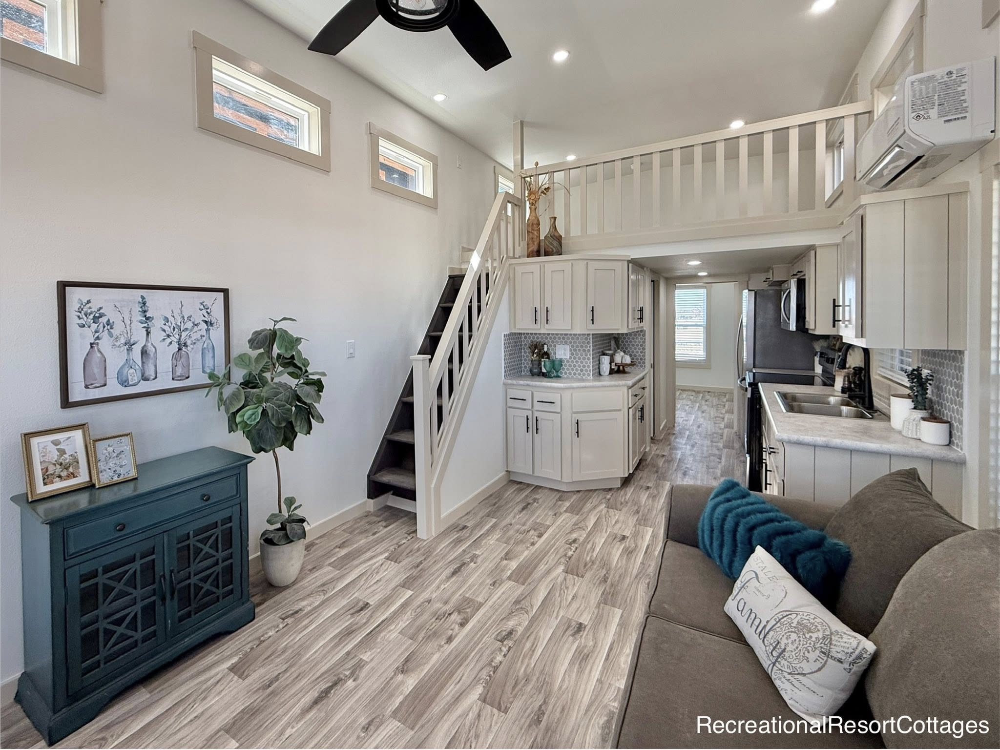 Bright, modern tiny house interior with wood flooring, a loft, and stairs. Features a compact kitchen, teal accents, cozy couch, and leafy decor.