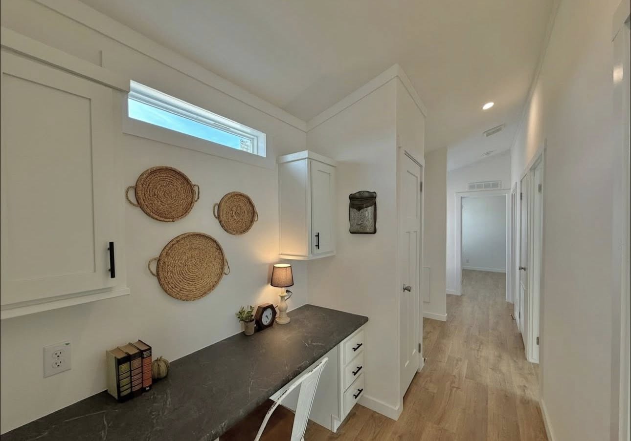 A cozy, well-lit hallway workspace features a black desk with books and a lamp, wicker baskets on the wall, white cabinets, and a bright wooden floor.