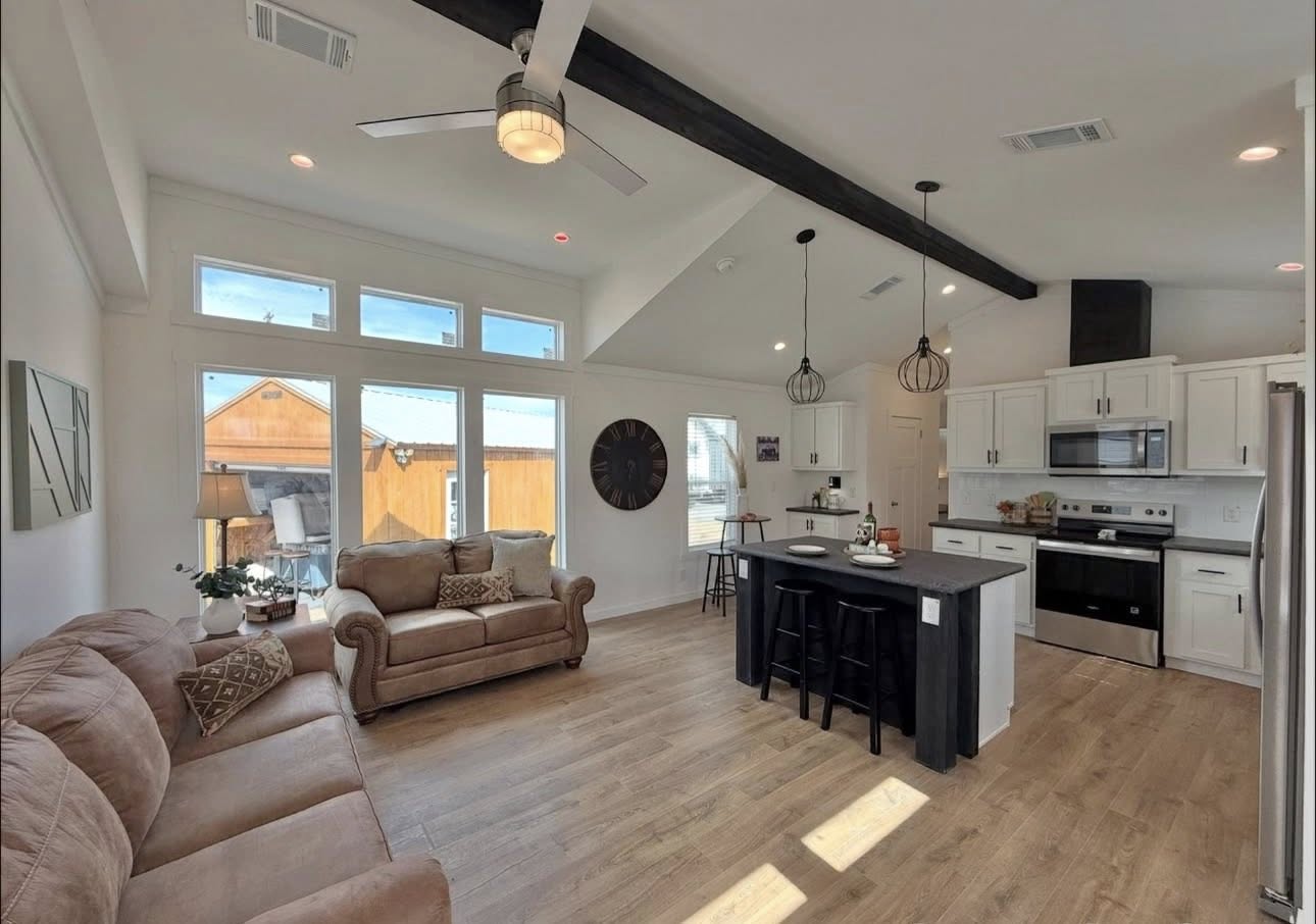 Modern open-concept living room and kitchen with high ceilings. Features cozy brown sofas, a black kitchen island with bar stools, and abundant natural light.