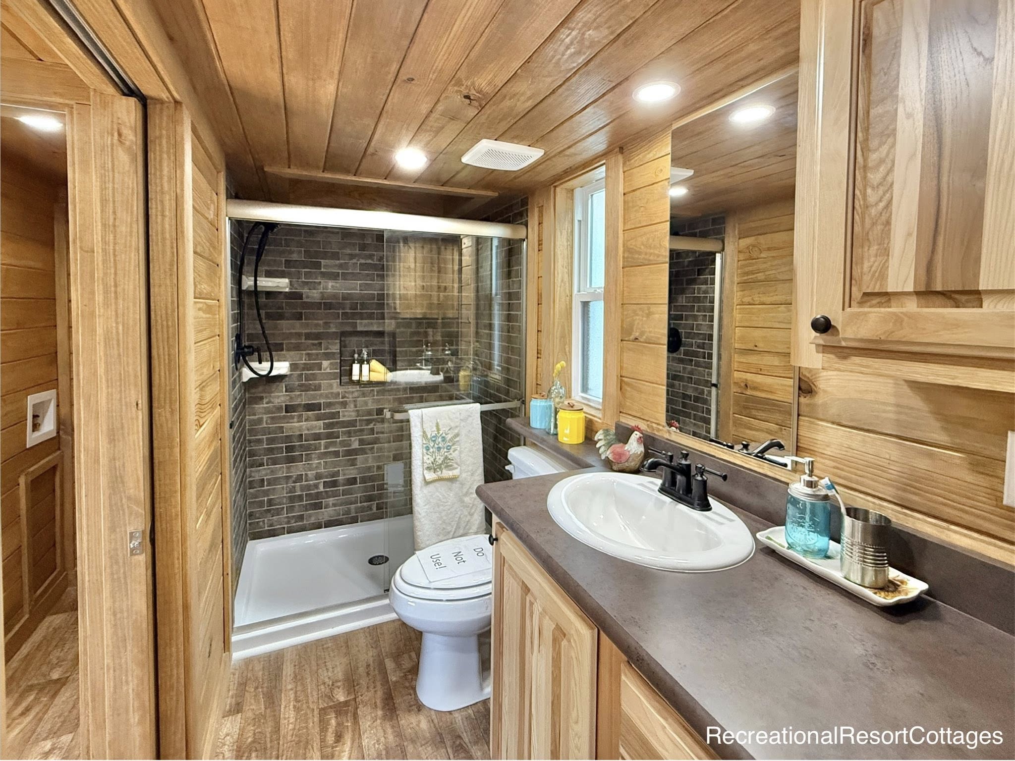 Warm, rustic bathroom with wood paneling, stone shower tiles, and a white sink on a brown countertop. Soft lighting creates a cozy atmosphere.