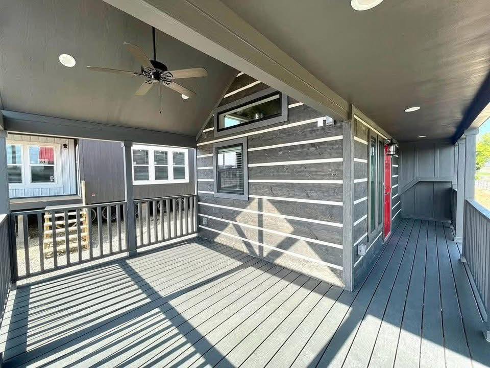 Covered porch with a dark gray wooden deck, fan on the ceiling, and a bright red door on the right. Sunlight creates a striped shadow pattern. Calm 