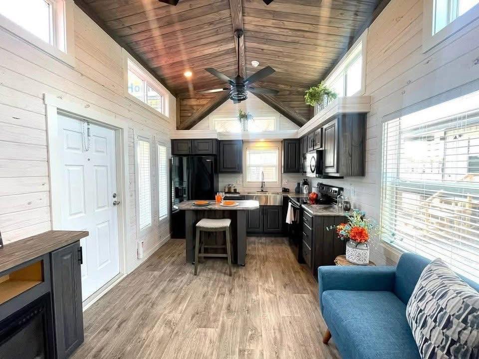 Cozy kitchen-living area with vaulted wood ceiling, dark cabinets, and modern appliances. Blue couch, small dining island, bright and inviting.