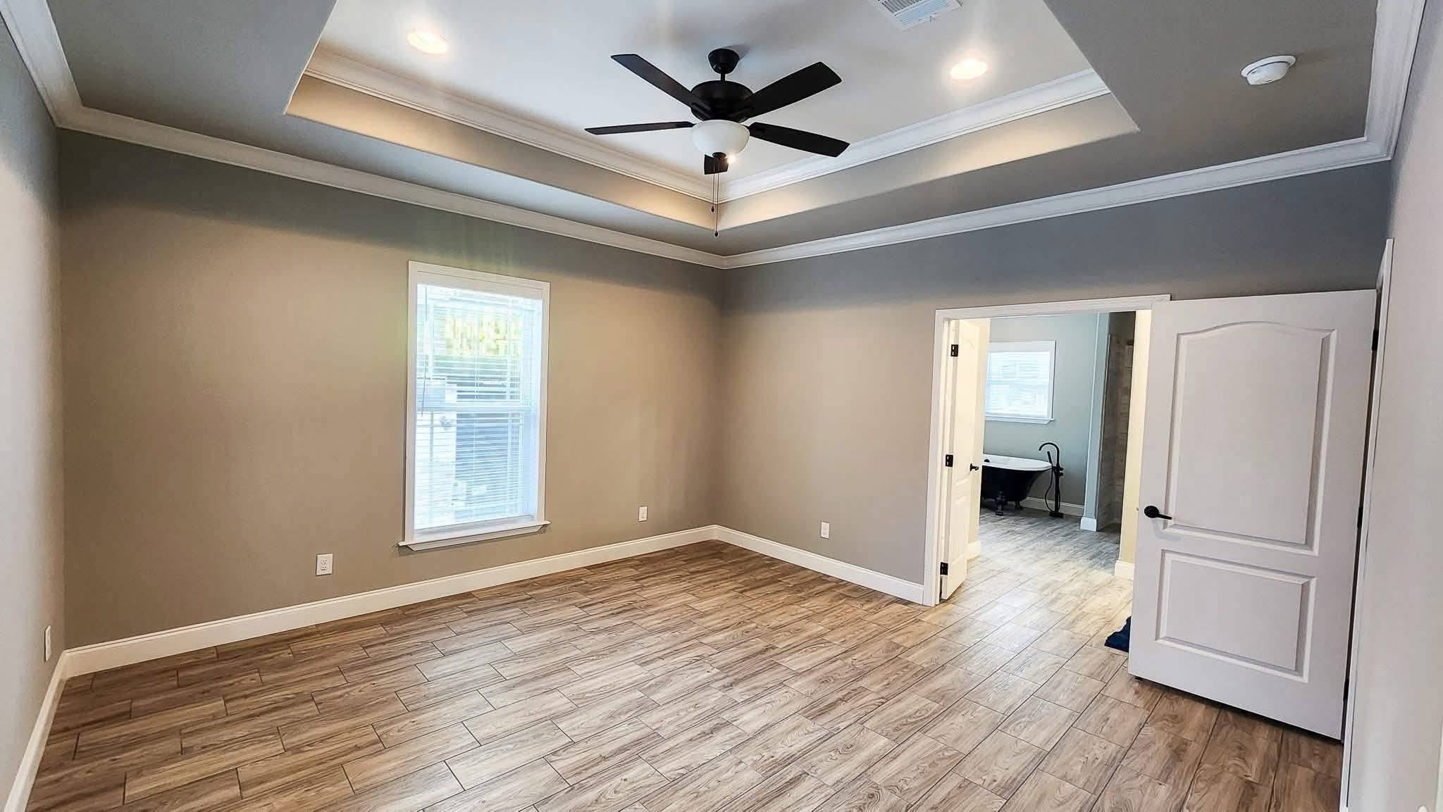 A spacious, empty room with light wood flooring, gray walls, and a tray ceiling. A window and ceiling fan provide natural light and modern ambiance.
