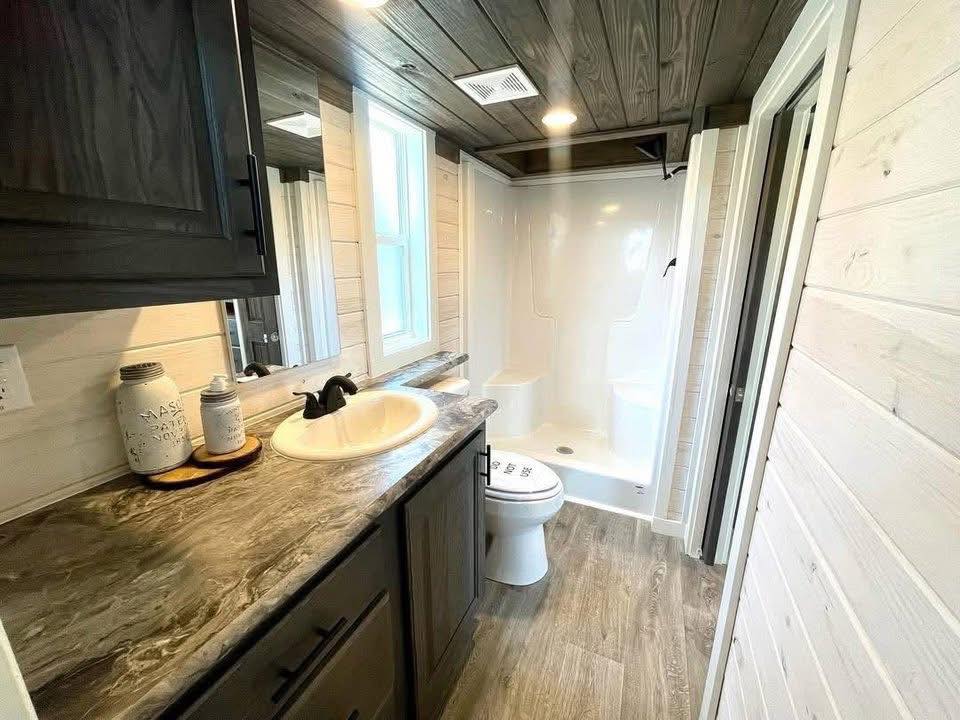 Cozy bathroom with rustic wood paneling, dark cabinets, and marble countertop. Features a sink, toilet, and spacious shower. Warm, inviting ambiance.