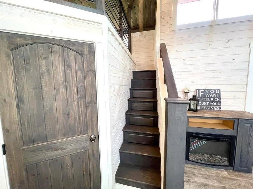 Cozy stairway with dark wooden steps and white walls. A wooden door to the left and a small fireplace beneath. A sign reads, "If you want to feel rich, count your blessings."