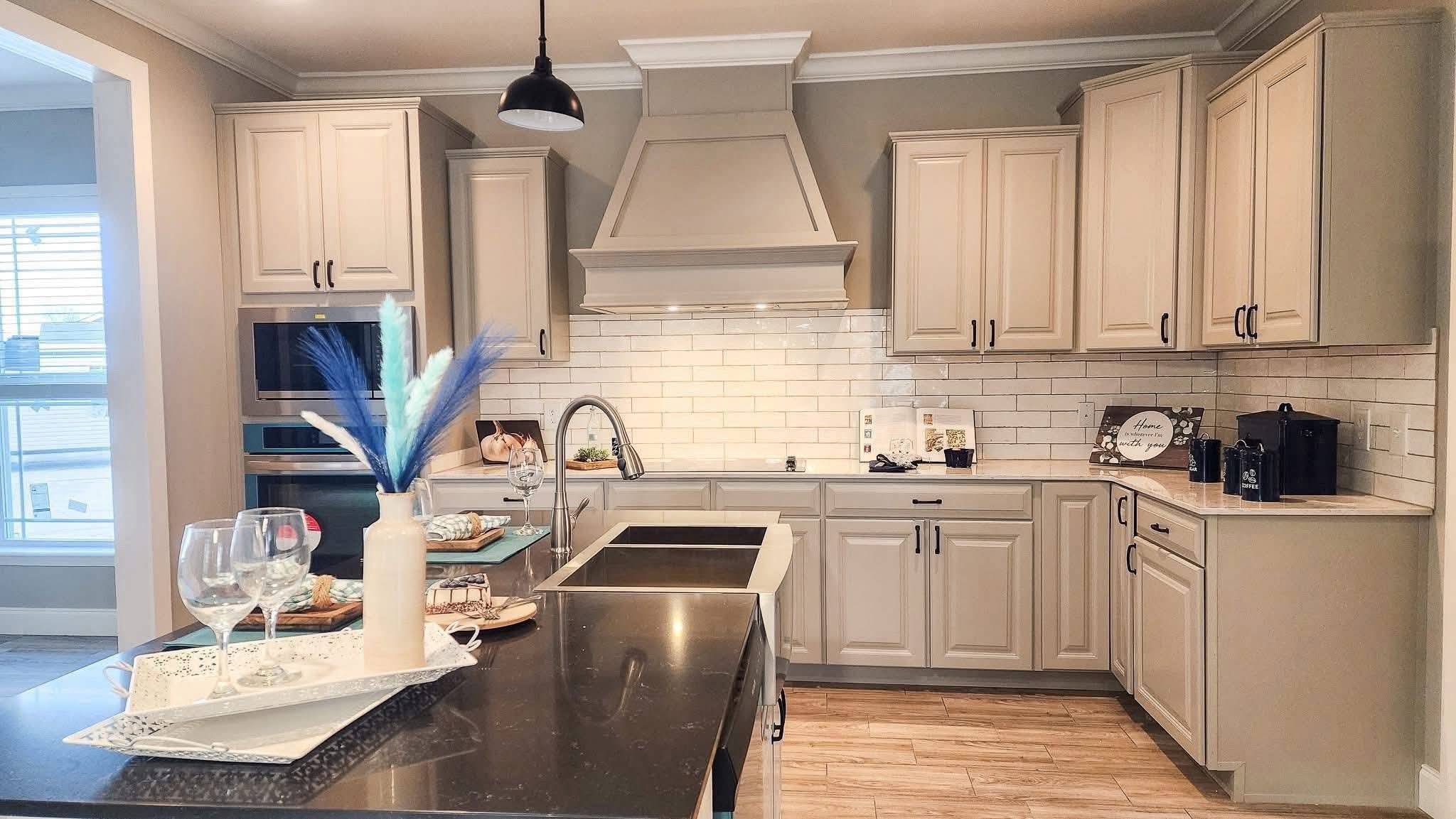 Modern kitchen with beige cabinets, a black countertop, and white subway tile backsplash. A vase with blue feathers and wine glasses adorn the island. Cozy and elegant.