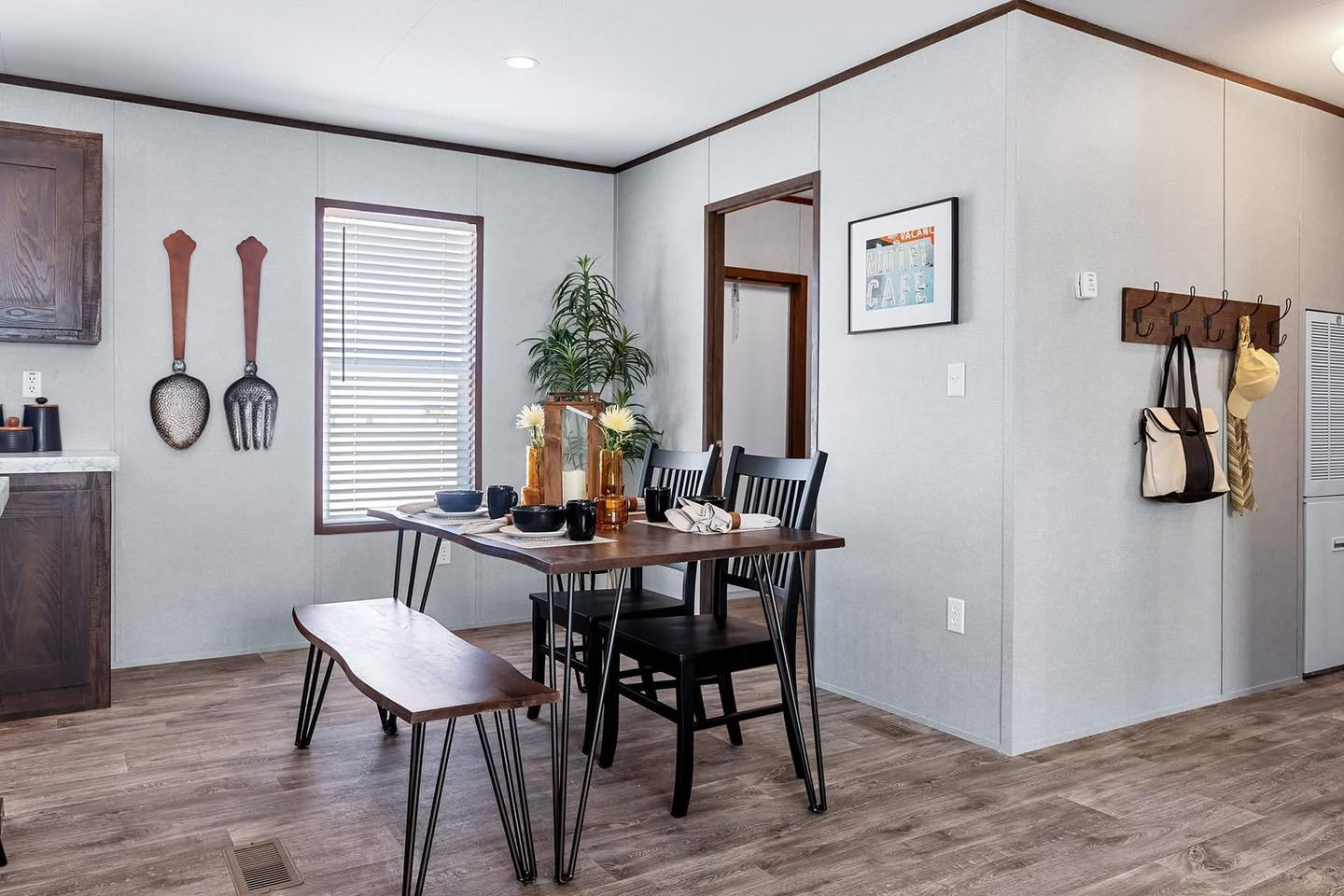 Modern dining area with a wooden table, black chairs, and a bench. Decor includes plants, wall art, and hanging utensils. Bright, cozy ambiance.