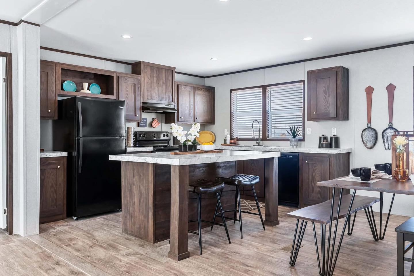 Modern kitchen with dark wood cabinets, black fridge, and marble countertops. Bar stools at an island. Natural light from a window creates a cozy feel.