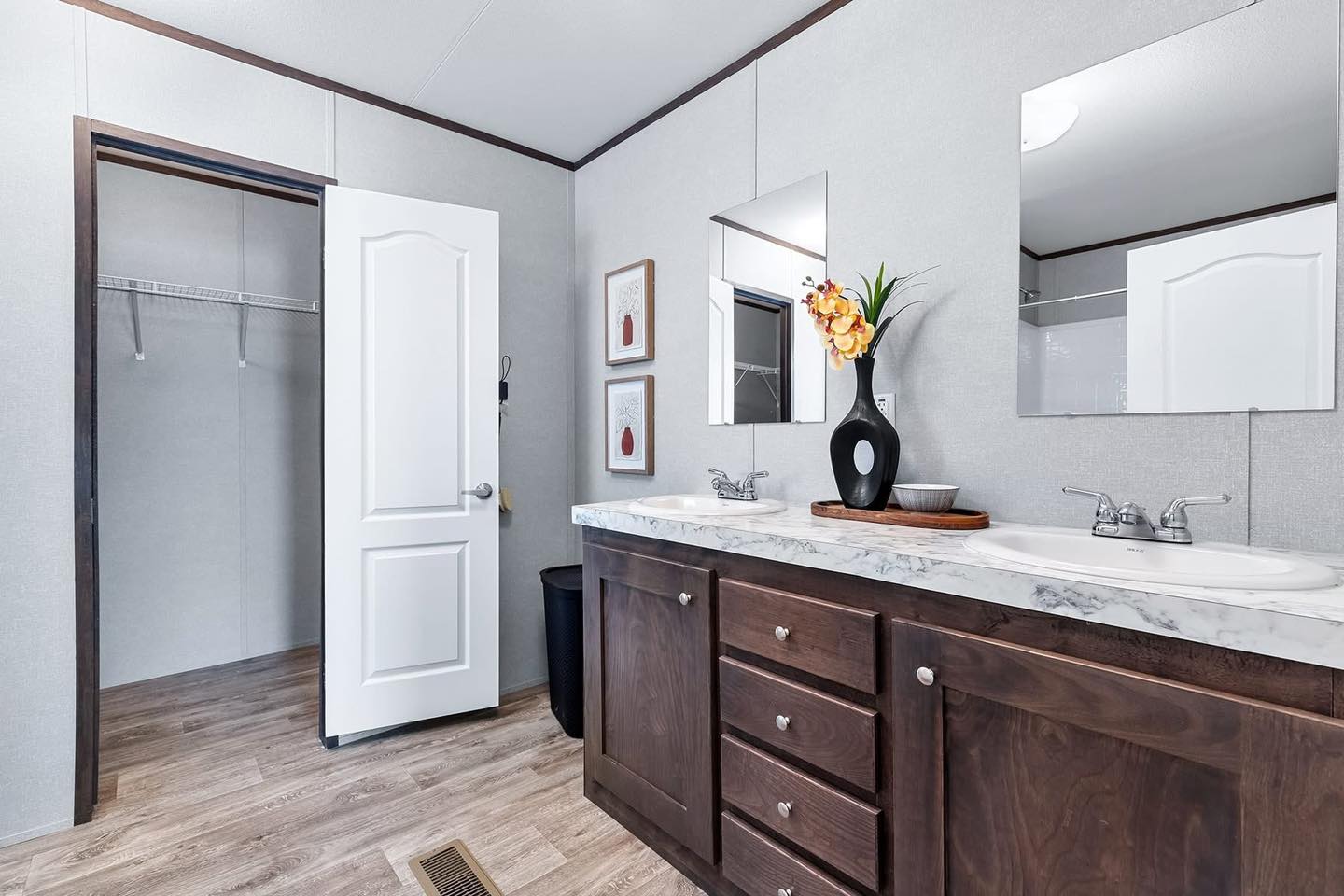 A modern bathroom with dark wood cabinets, a dual sink vanity with marble countertop, two mirrors, and a decorative vase. A door to a closet is open.