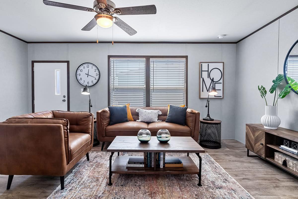 Cozy living room with brown leather sofas, a central coffee table, wall clock, minimalist decor, and a fan. Natural light from blinds.